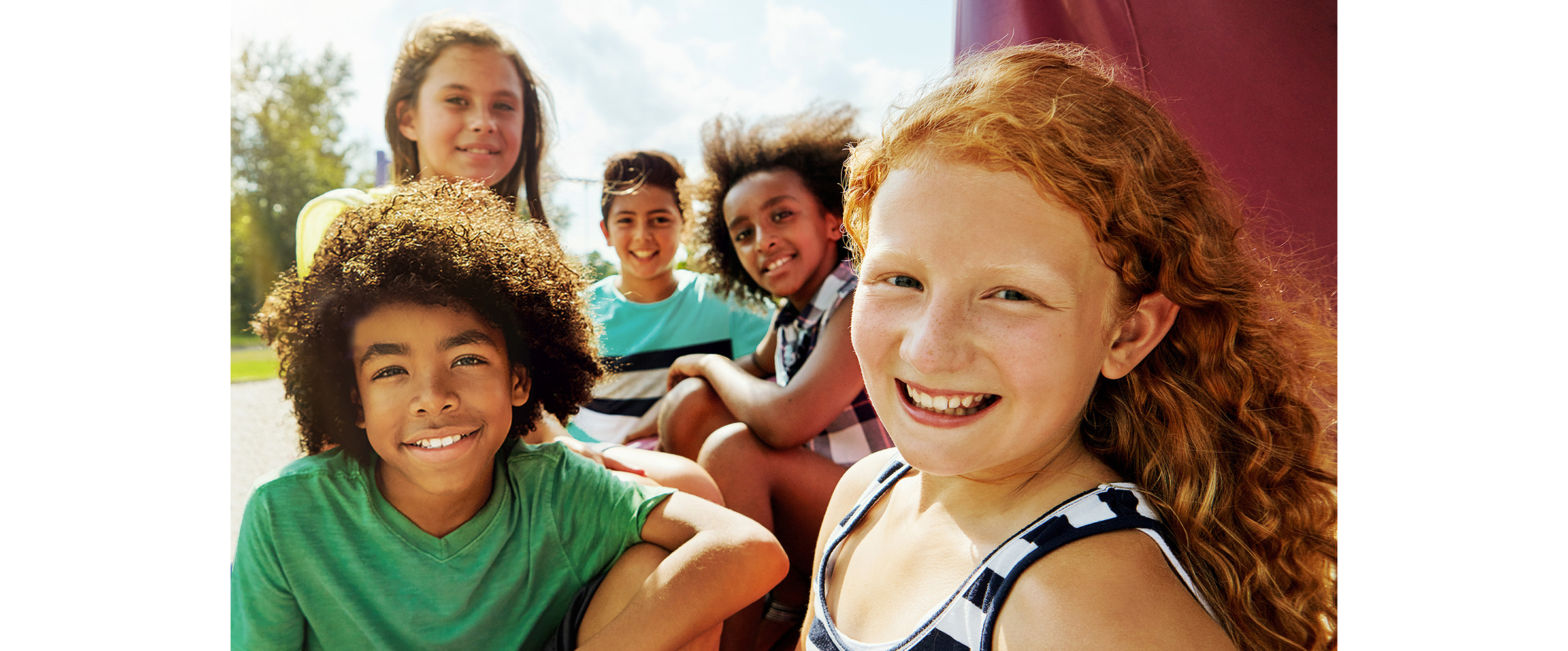Fünf Kinder sitzen im Freien und lächeln an einem sonnigen Tag in die Kamera. Im Hintergrund sind Bäume und blauer Himmel zu sehen.
