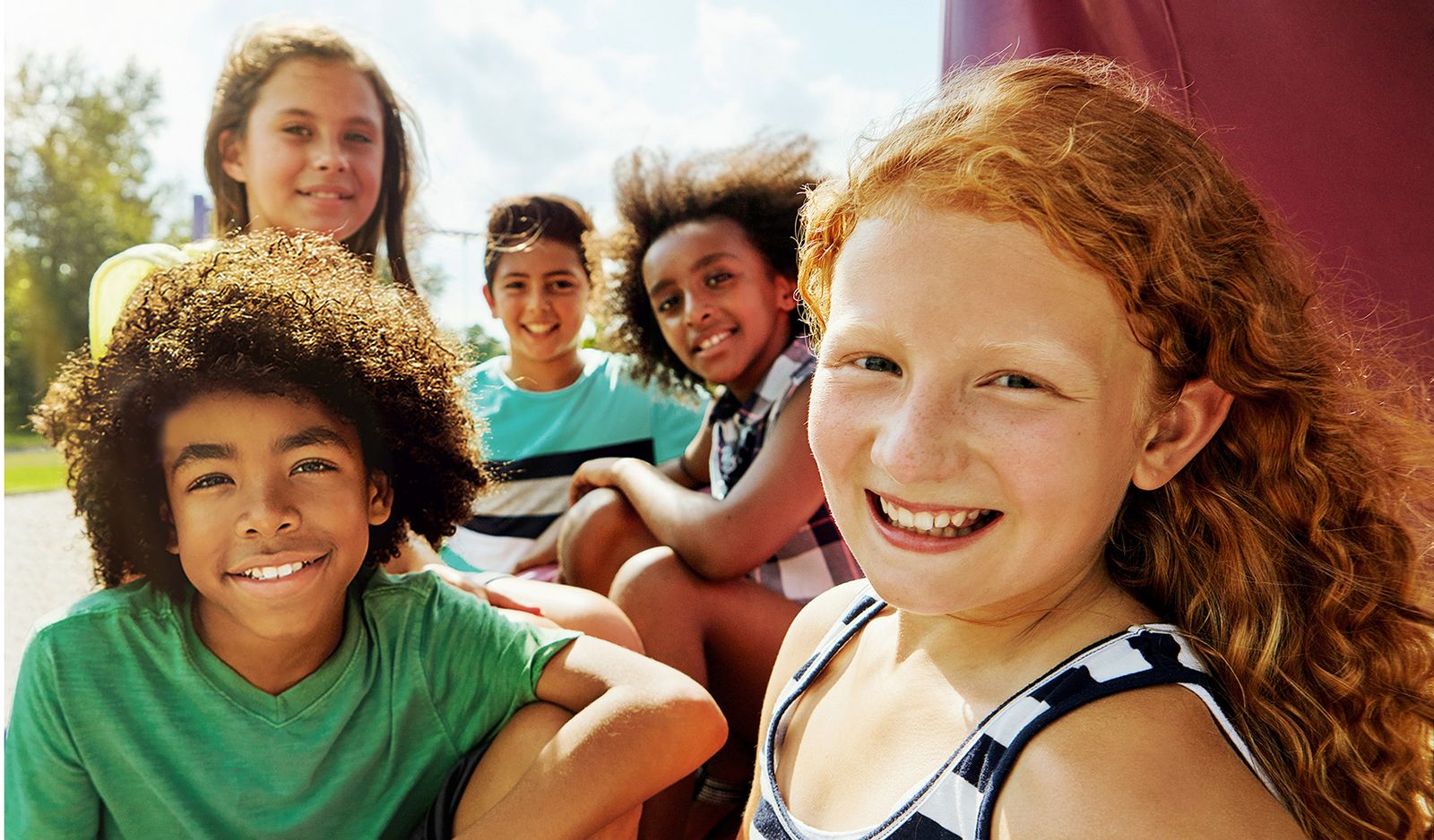 Fünf Kinder sitzen im Freien und lächeln an einem sonnigen Tag in die Kamera. Im Hintergrund sind Bäume und blauer Himmel zu sehen.
